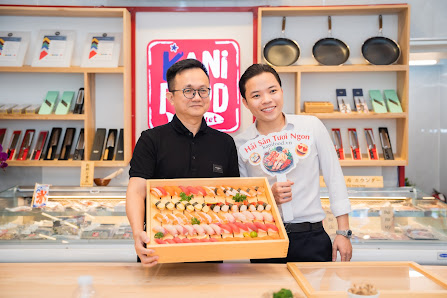Two individuals are standing in a food establishment holding up a box of sushi rolls and sashimi on display at the counter.