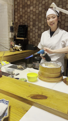 A person in a chef's uniform is using a blowtorch on food items placed on wooden trays at what appears to be an indoor kitchen or cooking station.