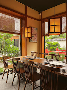 A traditional Vietnamese-style dining room with wooden tables and chairs, set for meals with plates and utensils. Large windows with bamboo blinds off