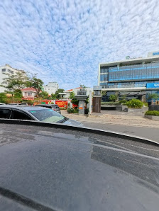 The image shows a view from inside a vehicle looking out onto an urban street scene with modern buildings and greenery under a partly cloudy sky.
