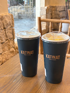 Two black cups labeled 'KATINAT' sit on a wooden table in what appears to be an indoor setting with stone walls and large windows, suggesting it could
