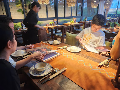 A restaurant interior in Thao Dien, Saigon, featuring a wooden table with diners. There are plates, chopsticks, a tablet, and a person reading. A serv