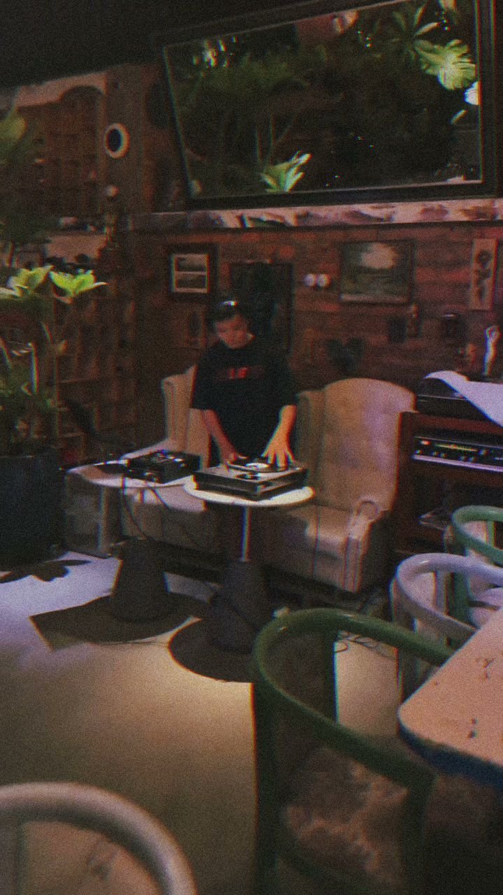 A person is standing in a dimly lit room that appears to be set up for DJing or music production, with various electronic equipment and plants visible