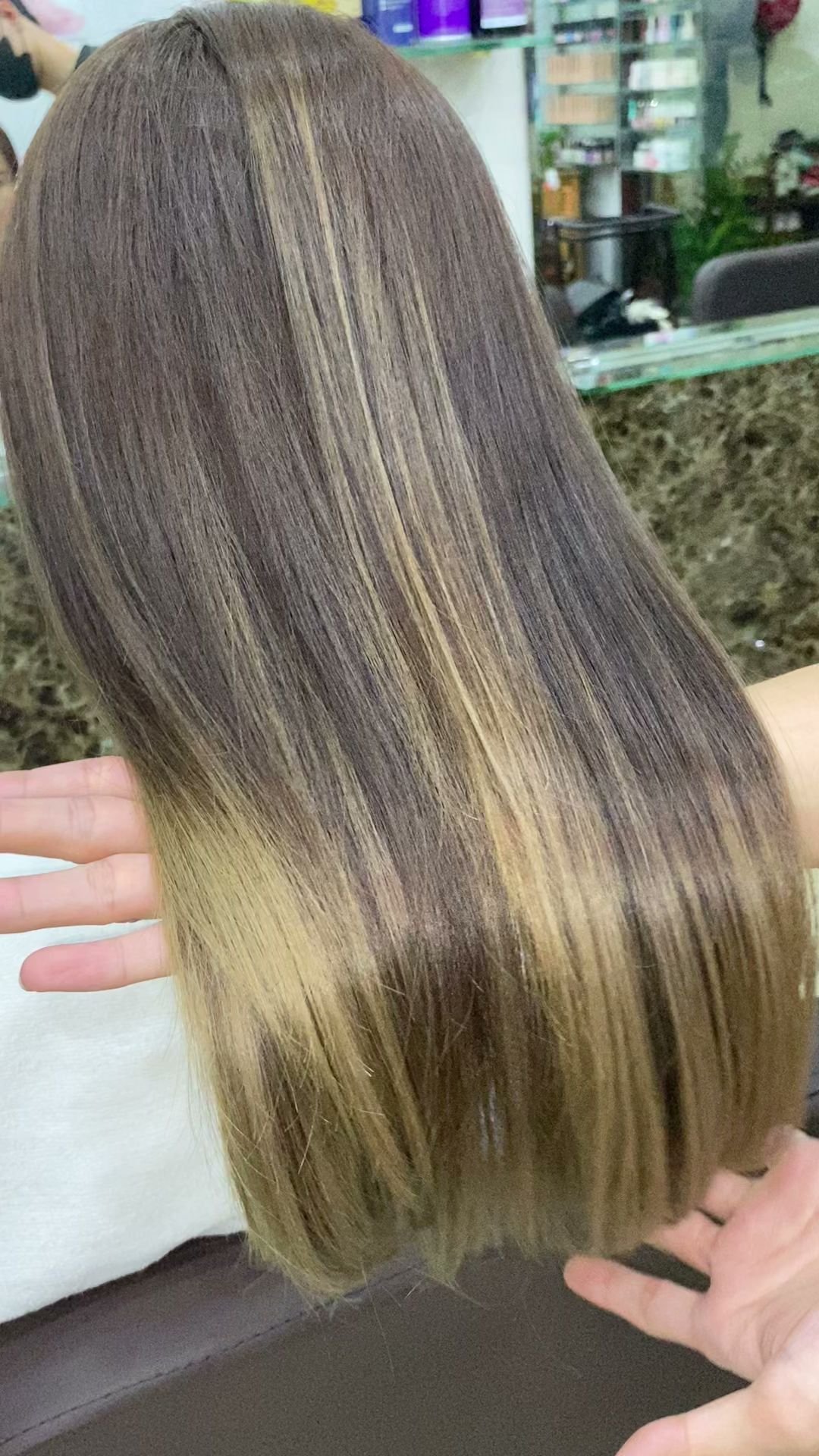 A close-up of a person's hair in what appears to be a salon setting with shelves stocked with products visible in the background.