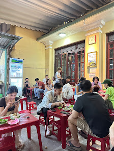 A bustling restaurant interior in Thao Dien, Saigon, featuring patrons seated at red plastic tables and stools. The space has yellow walls, large wind
