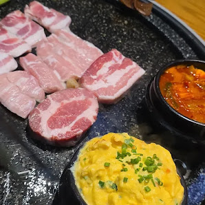 The image shows a variety of Korean dishes including sliced pork belly (samgyeopsal), kimchi in a black bowl, and a side dish that appears to be seaso