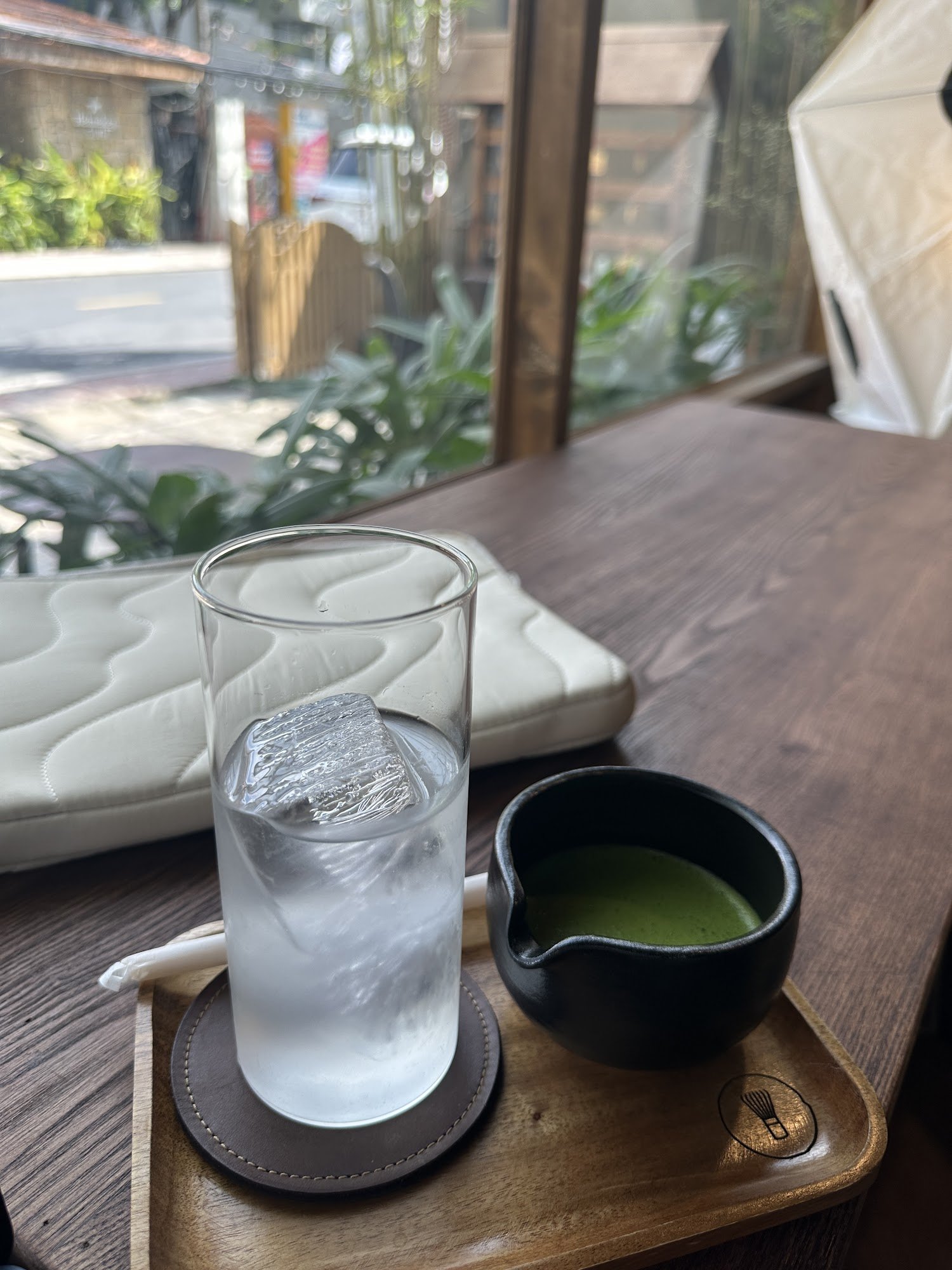 A glass of water with a large ice cube sits on a wooden tray alongside a bowl containing green tea in an indoor setting, possibly a cafe or restaurant
