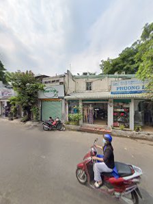 An exterior street scene in Thao Dien, Saigon featuring a motorbike with a rider, various storefronts including one with a 'PHUONG' sign, trees, and a