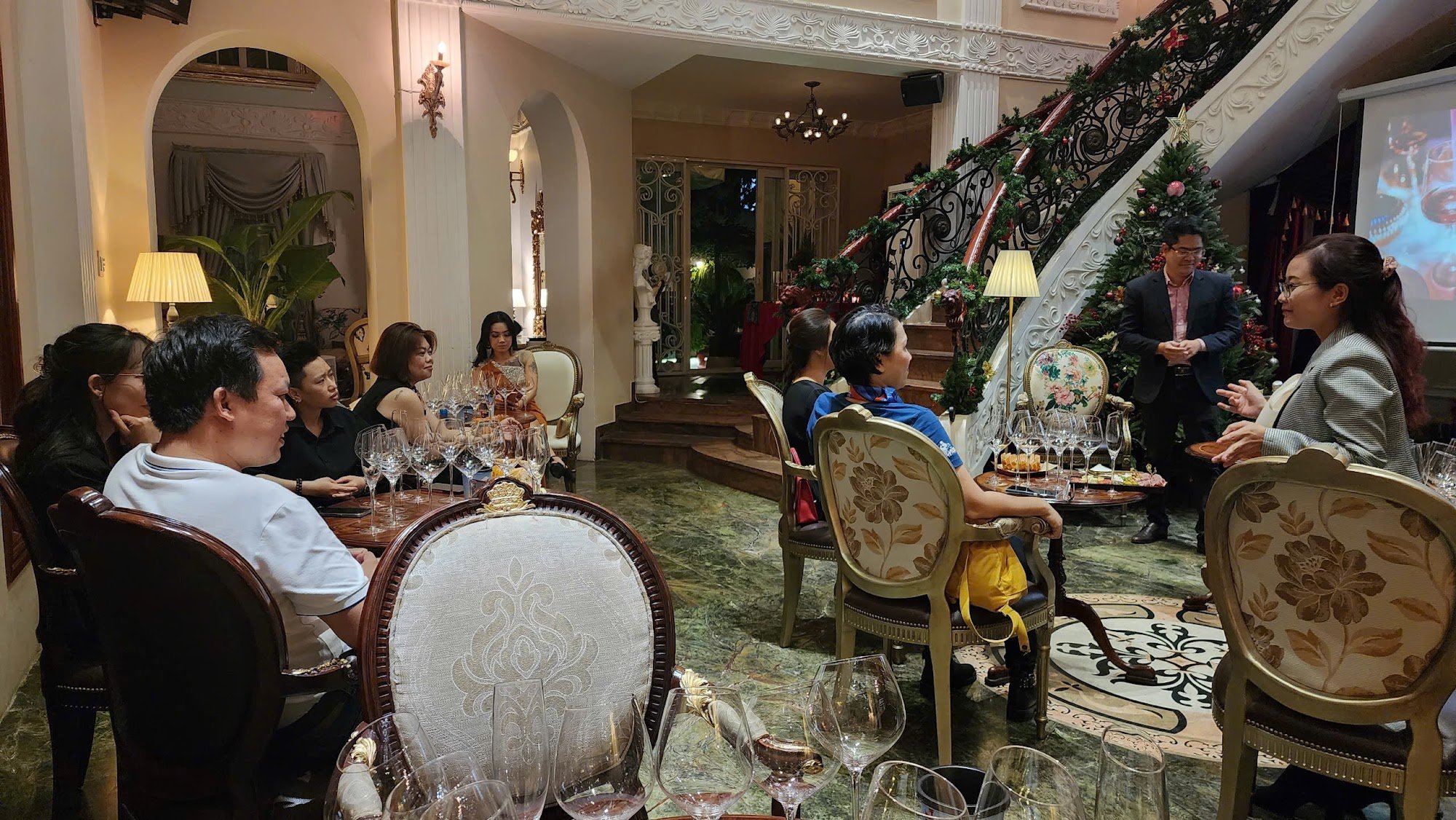 A formal indoor gathering in a luxurious venue with ornate decor. People are seated around tables with glassware, some standing near a staircase adorn