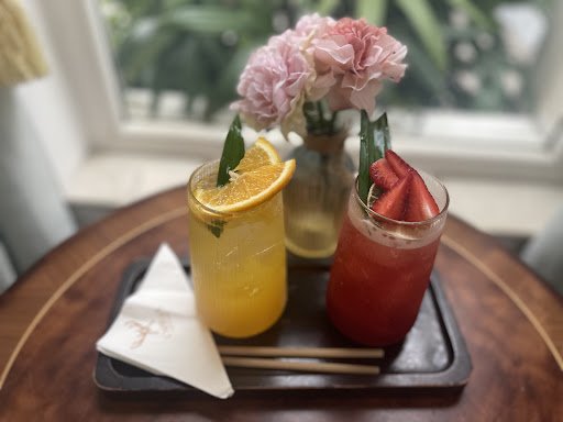 The image shows a wooden tray holding two colorful cocktails and a vase of pink flowers in the background.