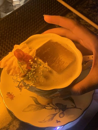 A close-up of a hand holding a cup containing what appears to be an alcoholic beverage with ice cubes and garnished with edible flowers, served on a d
