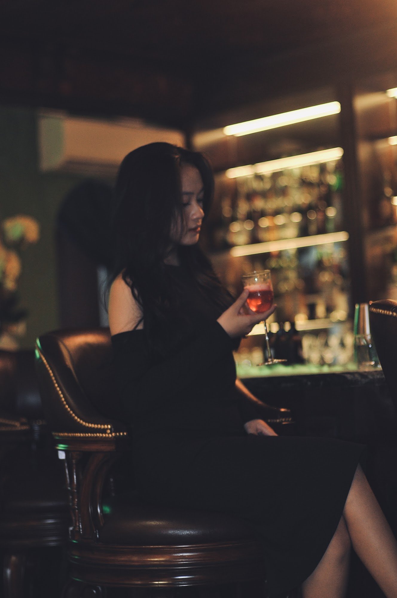 A woman with long dark hair, wearing a black off-shoulder dress, sits on a brown leather bar stool in a dimly lit bar. She holds a glass of red cockta