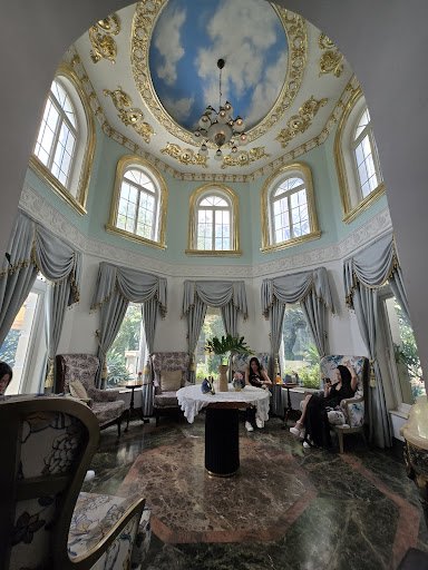 The image depicts an elegant and luxurious interior space featuring a high ceiling adorned with gold accents and blue sky motifs. Large windows allow 