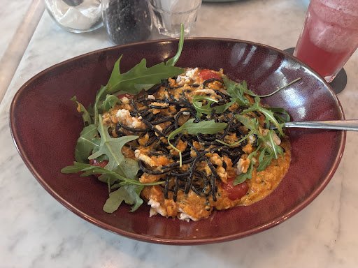 A bowl of food containing a mix of greens and what appears to be noodles or rice with black seaweed strips on top, served in a red sauce. The dish is 