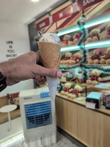 A hand holding a cone of ice cream in an indoor setting that appears to be the interior of a dessert shop or cafe, with various desserts displayed on 