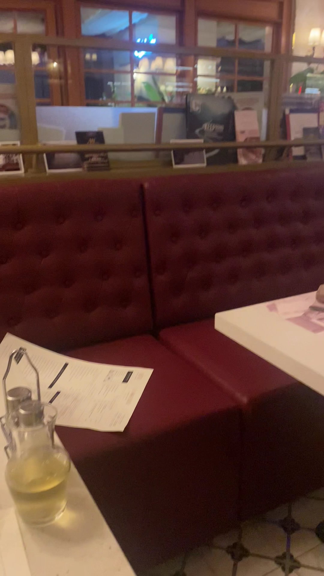 The image shows a cozy corner of an indoor venue featuring red tufted booth seating with a glass table in the foreground containing a pitcher and pape