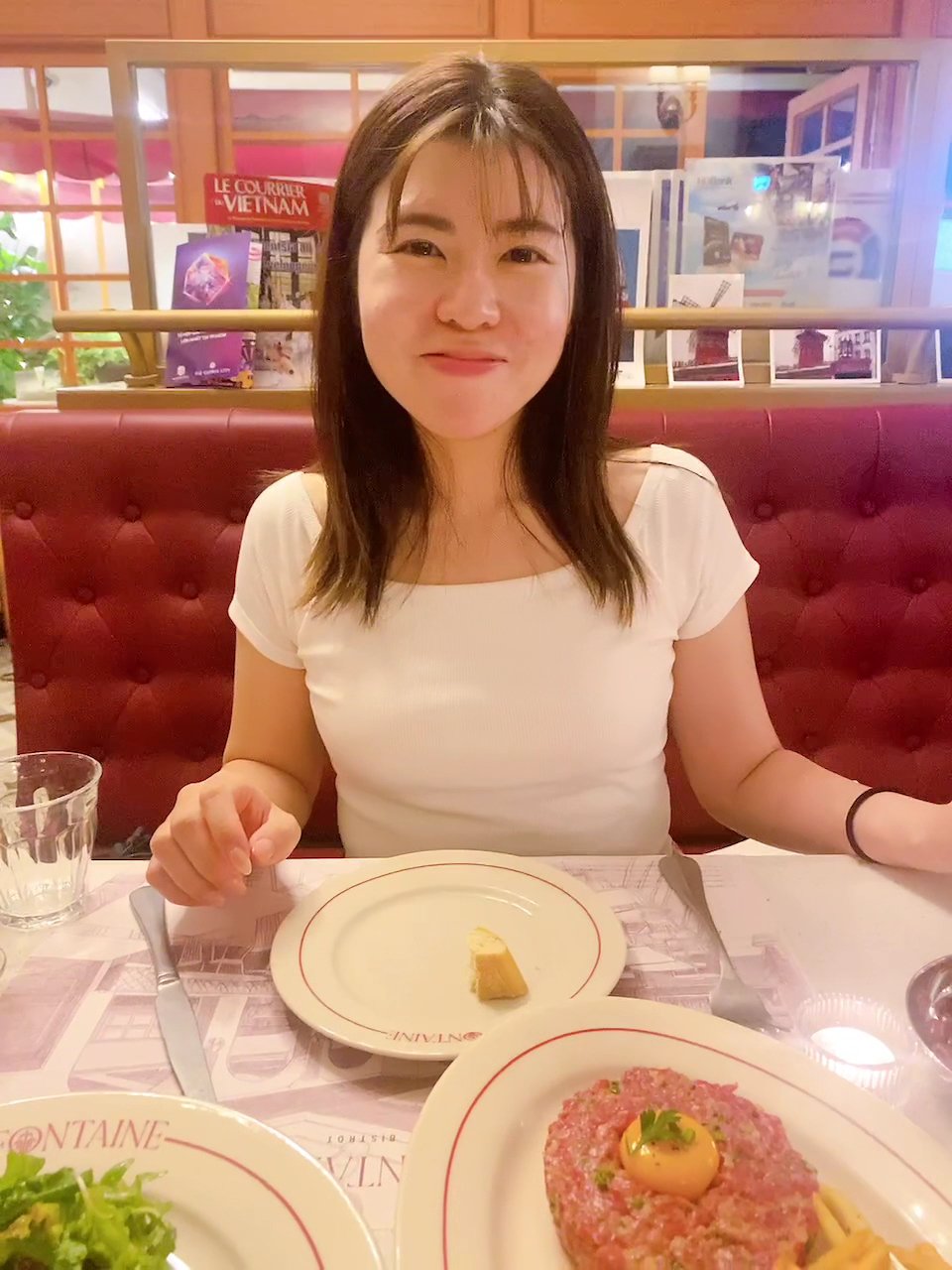 A woman is seated at a table in what appears to be an upscale restaurant or cafe with red upholstered seating and promotional materials visible on the