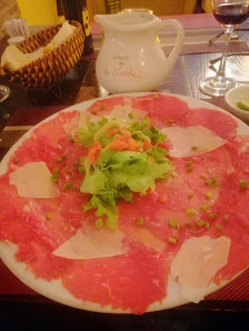 A plate of thinly sliced raw meat garnished with fresh greens and vegetables on a table in what appears to be an indoor dining setting.