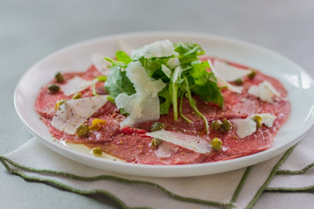 A plated dish featuring a pink-colored raw meat preparation garnished with greens and shaved cheese on top.