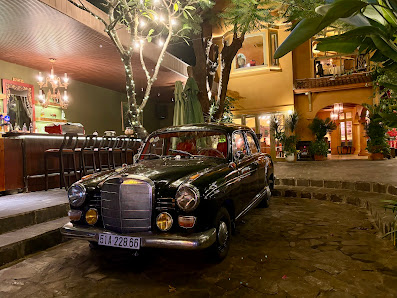 A nighttime exterior shot of a venue in Thao Dien, Saigon, featuring a classic black Mercedes-Benz car parked on a stone-paved area. The background in