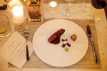 A plated dish consisting of a piece of meat on top of what appears to be mashed potatoes or puree, garnished with small vegetables and sauce.
