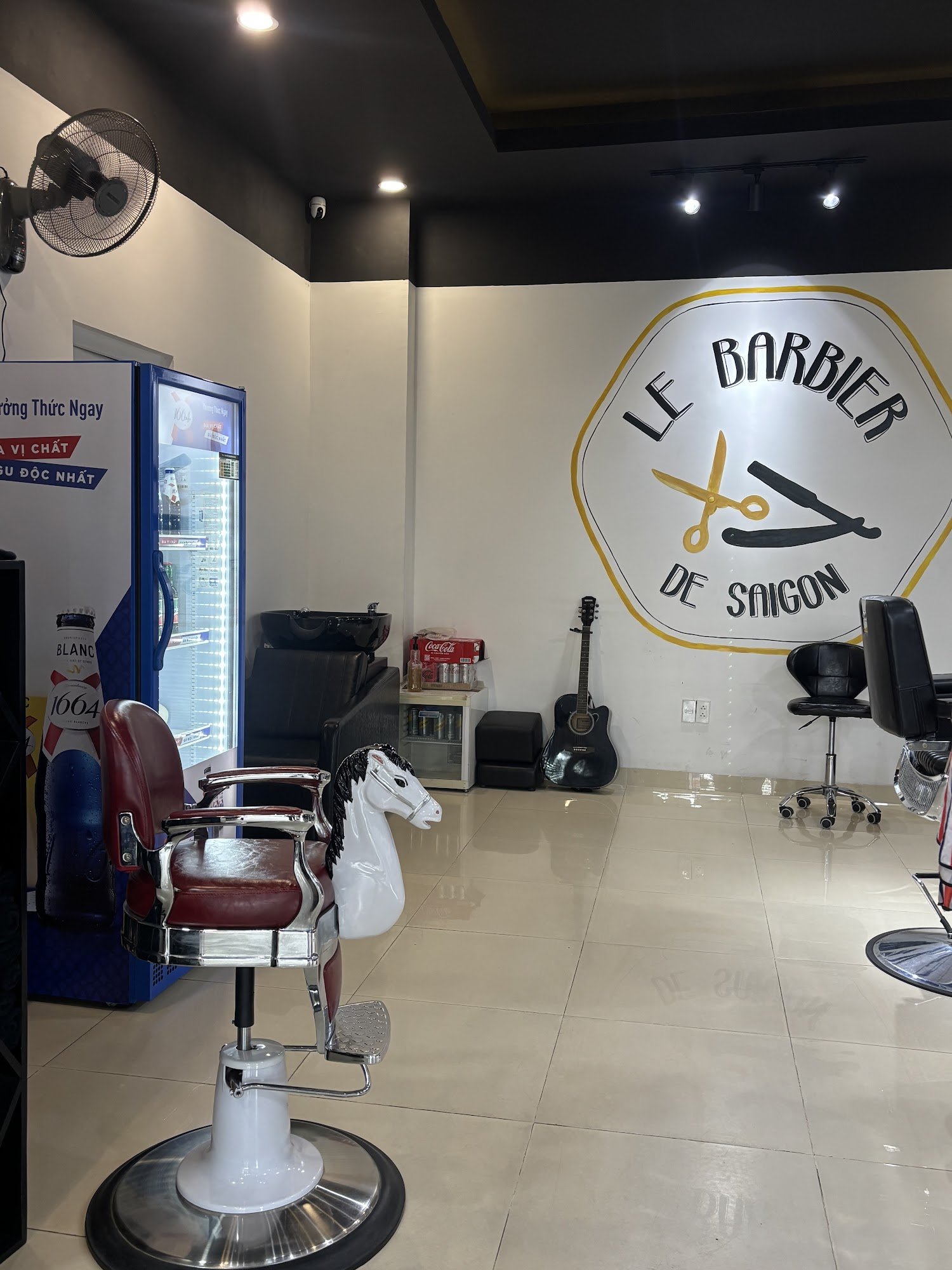 The image shows an interior view of a barber shop named 'Le Barbier de Saigon'. The space is clean and modern, with white tiled flooring and black wal