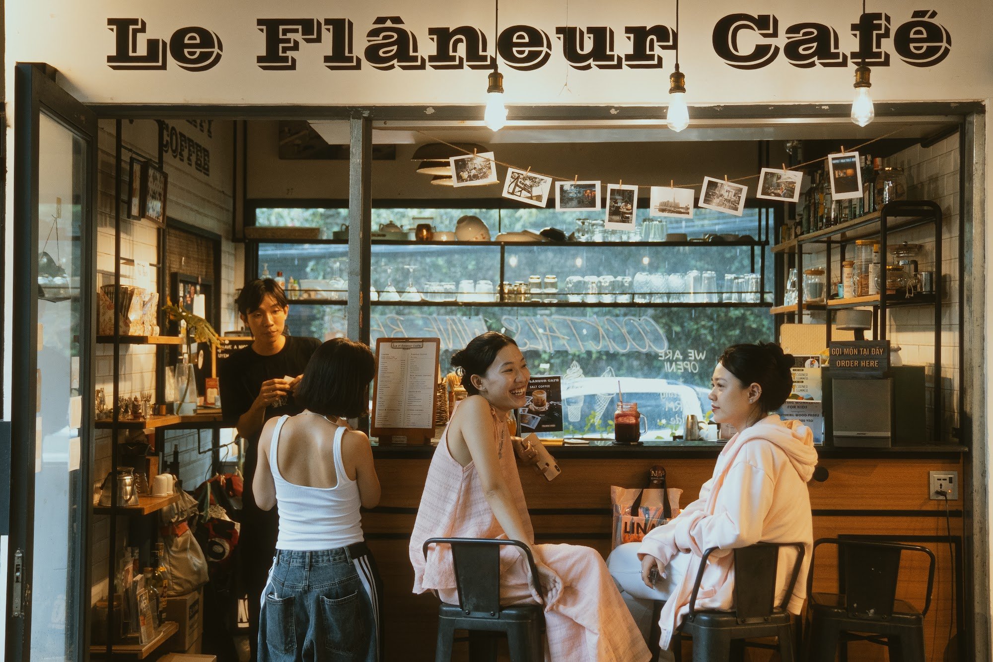 The image depicts an interior view of a café named 'Le Flanleur Café'. It shows three individuals engaged in conversation while seated at the counter,