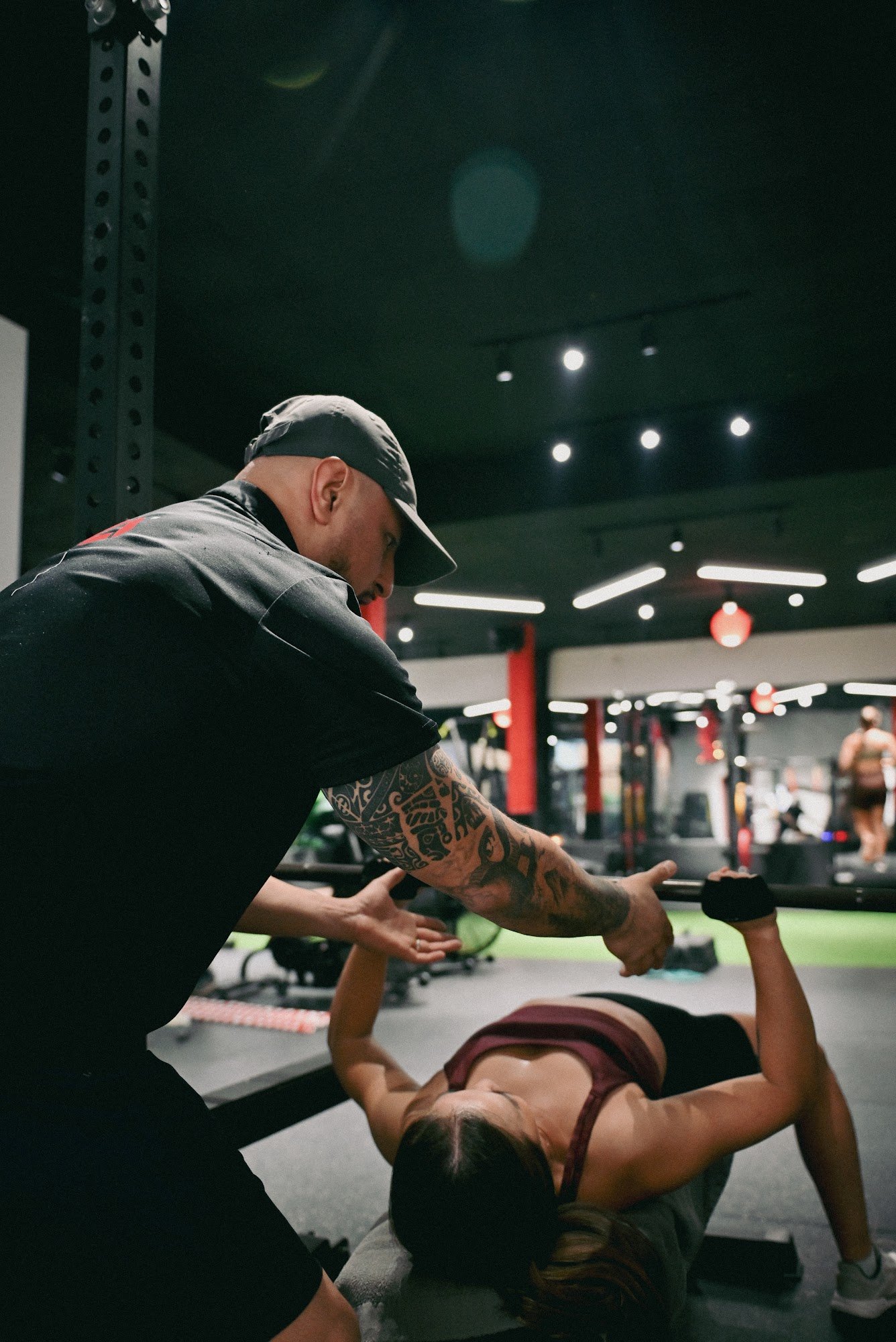 The image depicts an indoor gym setting where a personal trainer is assisting a female client with her exercise routine.