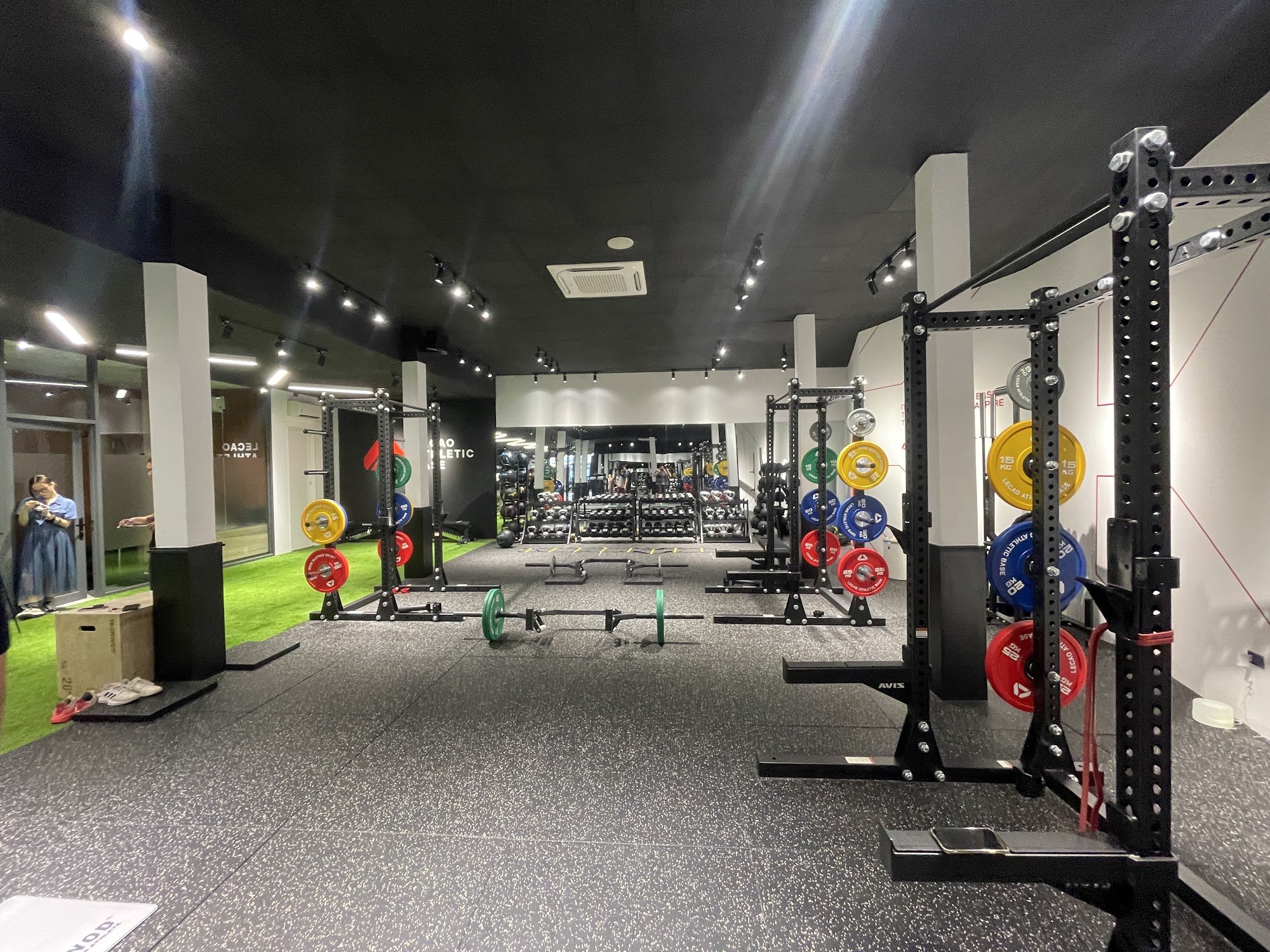 The image shows a well-equipped gym interior featuring weightlifting equipment such as barbells and plates on racks, squat stands with red weights, an