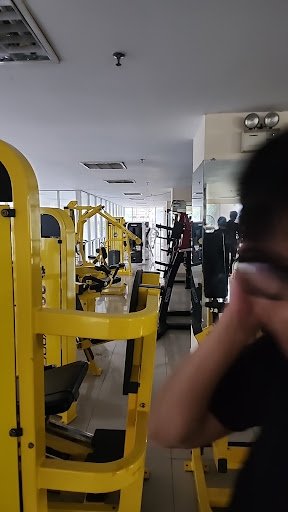 The image shows an interior view of a gym equipped with yellow exercise machines and various other fitness equipment in the background.