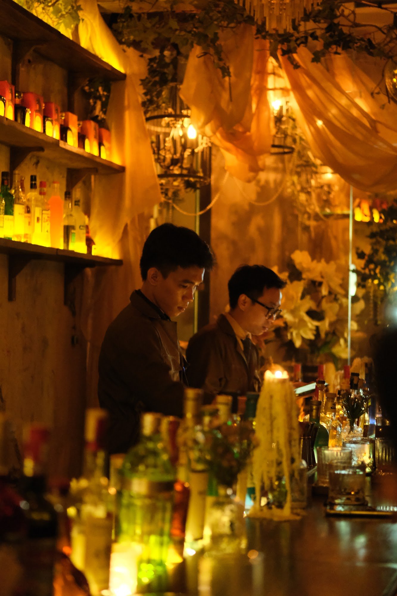 The image depicts the interior of a bar in Thao Dien, Saigon. Two staff members are working behind a wooden bar counter, surrounded by various bottles
