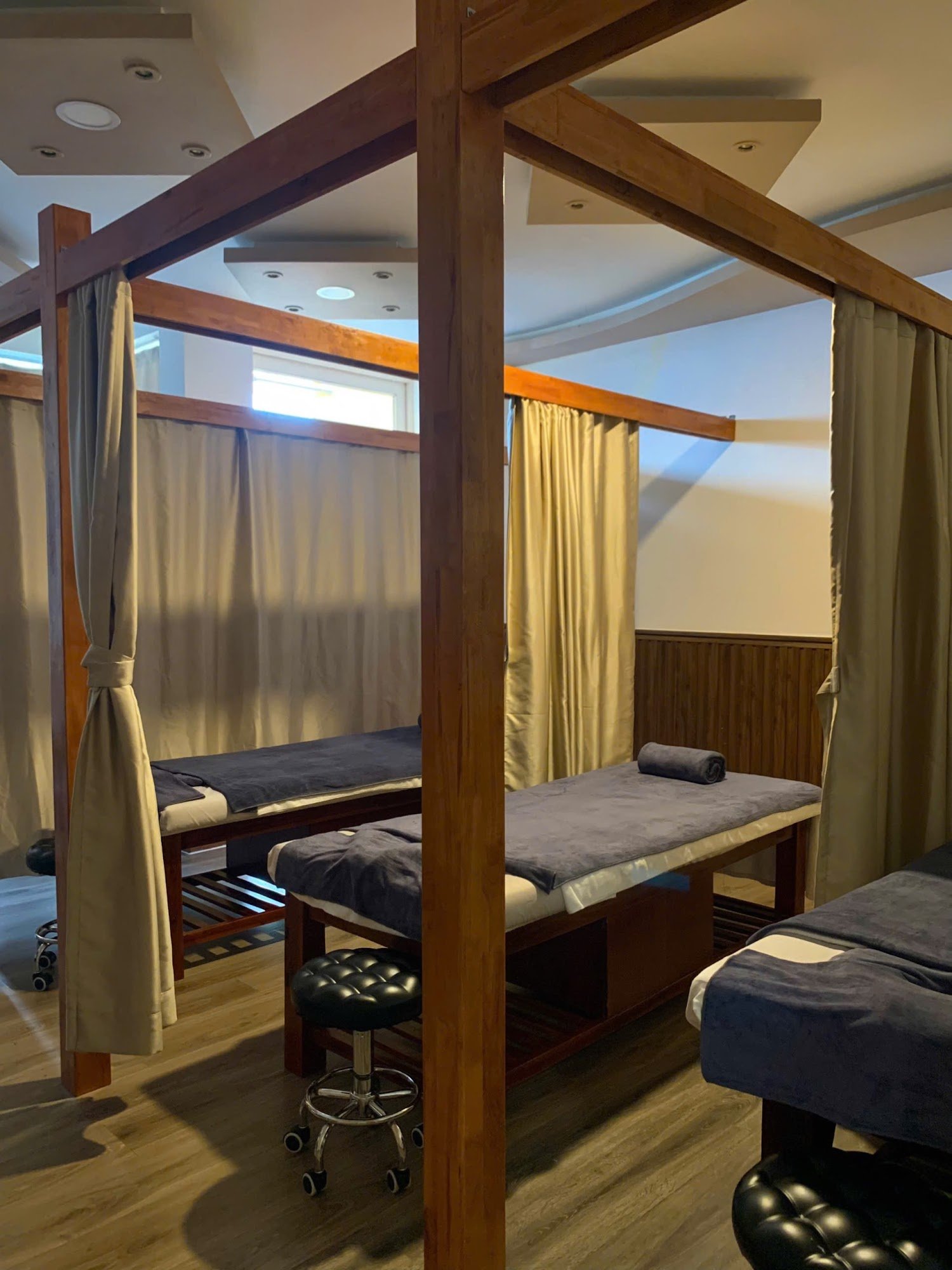 The image shows a well-lit room featuring two massage tables enclosed by wooden partitions with beige curtains on either side.