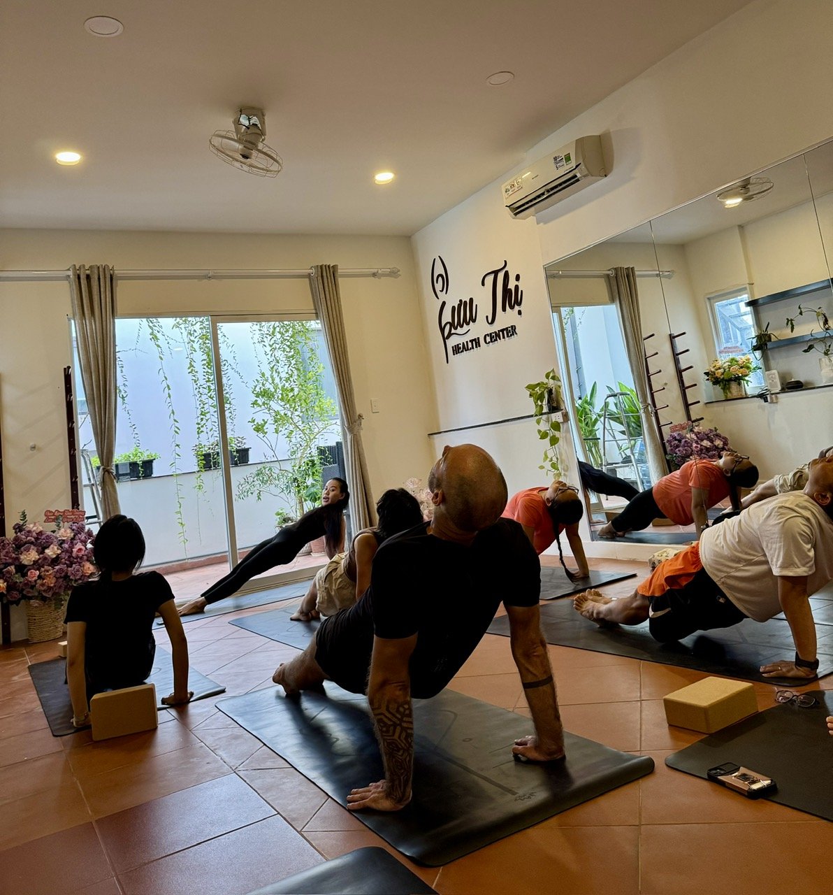A group of individuals is practicing yoga in a well-lit indoor space labeled 'Giai Thi Health Center'. The room features large mirrors, exercise mats