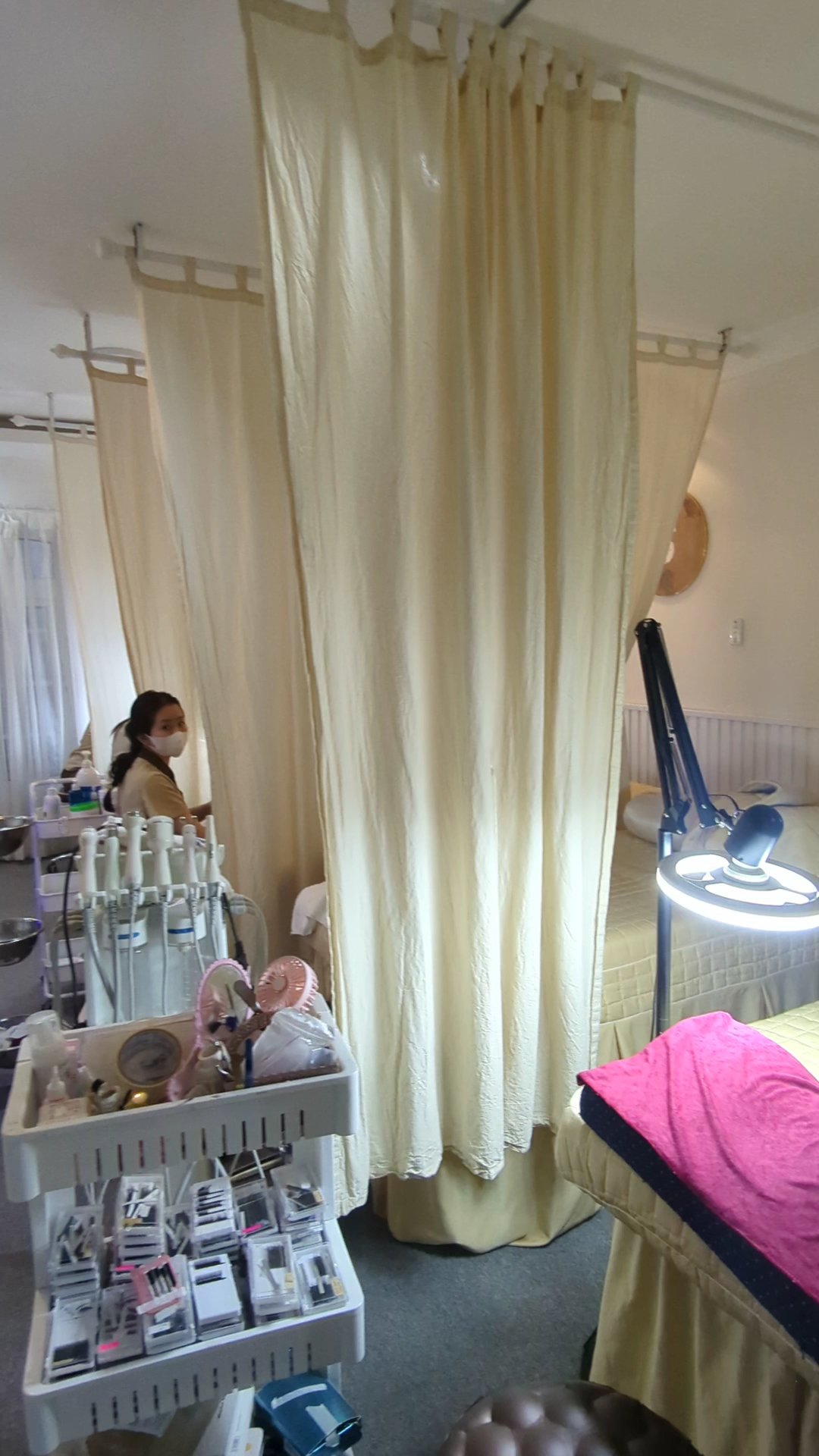 The image depicts an interior space that appears to be a medical or beauty treatment room with beige curtains and various equipment on shelves, includ