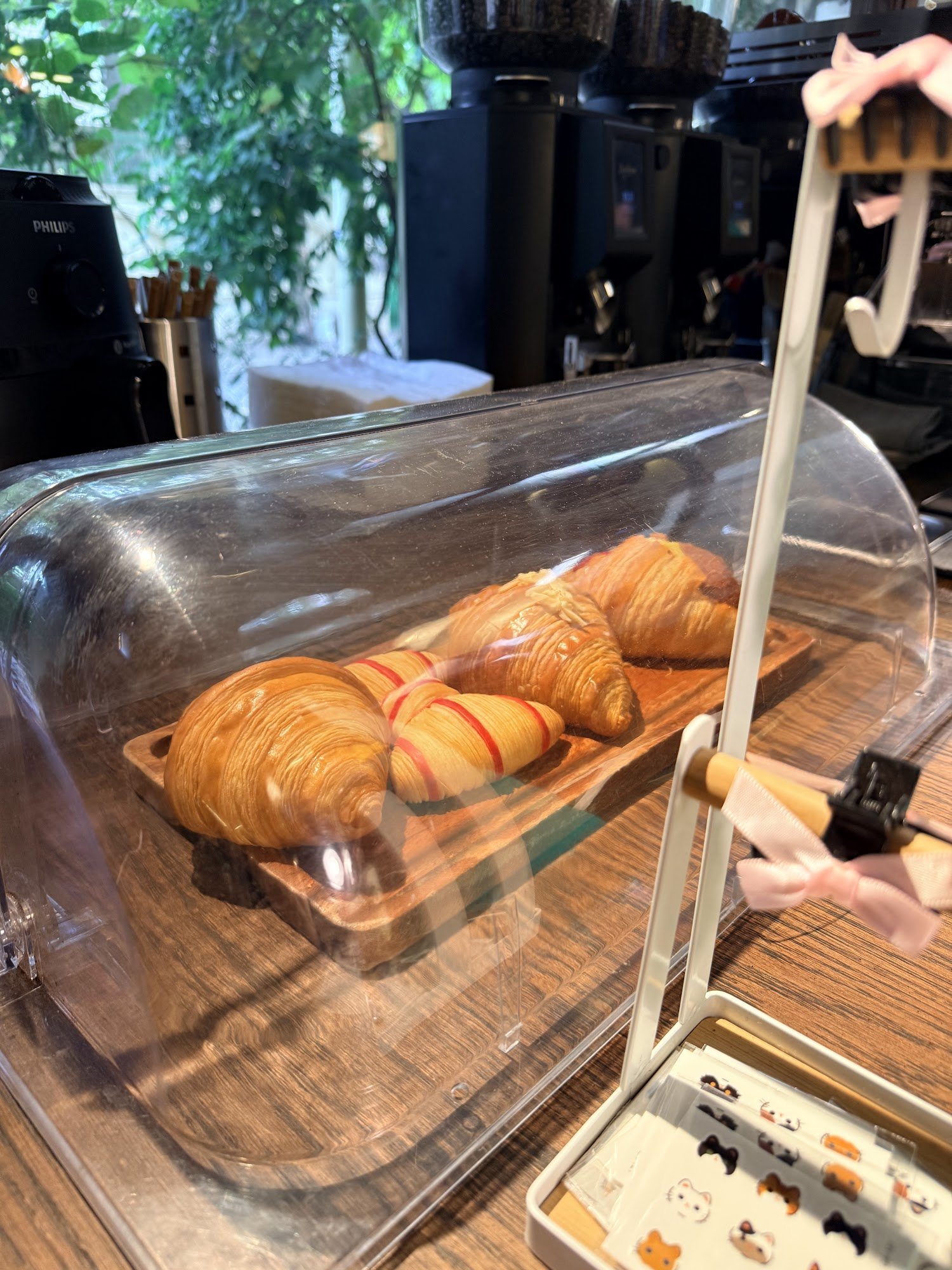 The image shows a display case in what appears to be a café or bakery setting with croissants on wooden boards inside the glass dome of the display ca