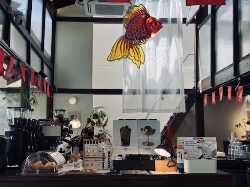 The image shows an interior view of a café or coffee shop with various items on display and decor elements such as plants, art pieces, and hanging dec