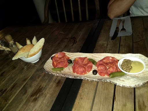 A wooden table set for a meal featuring an appetizer platter of sliced cured meats and olives on flatbread, accompanied by breadsticks in a bowl with
