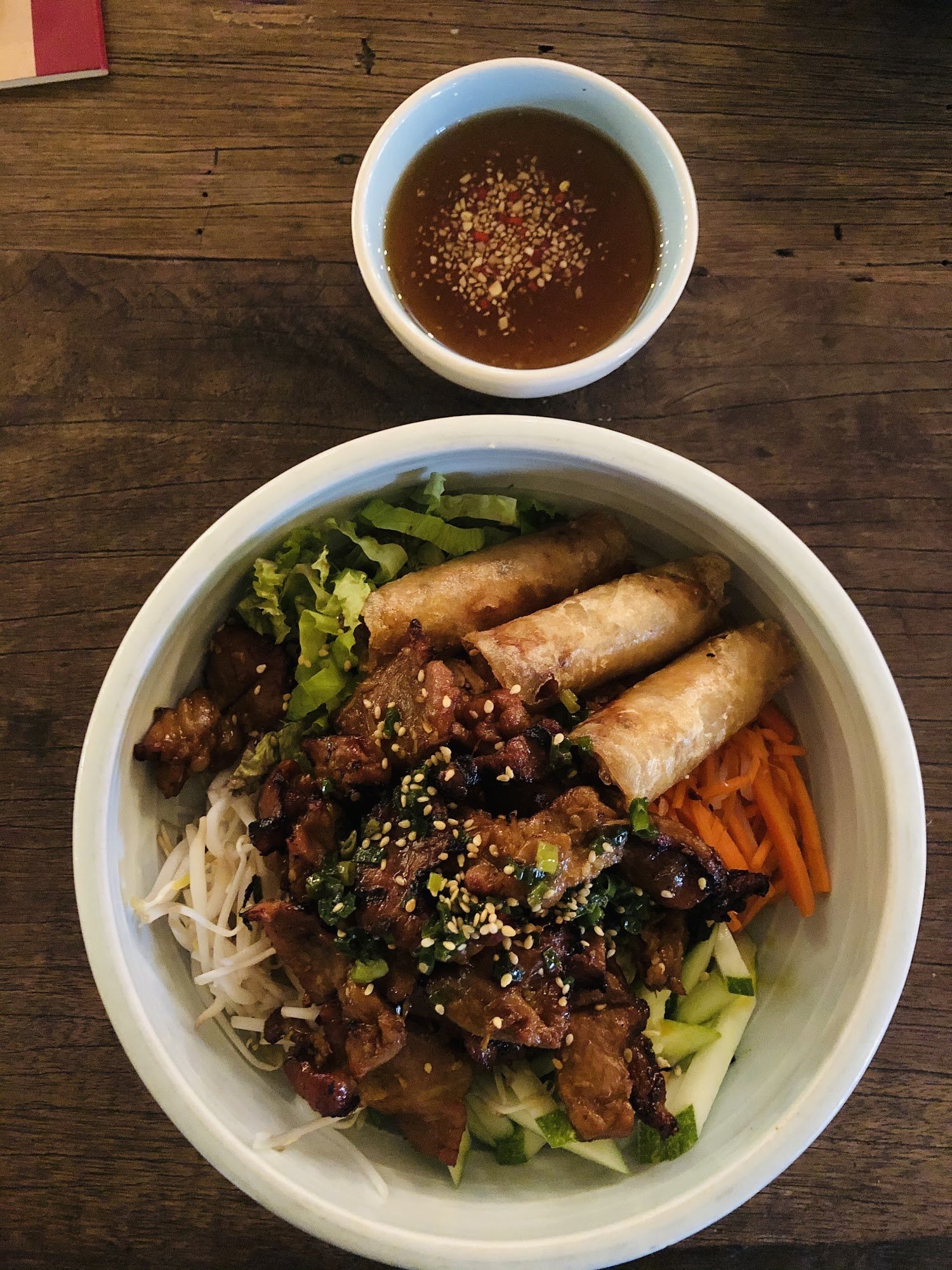 A bowl of Vietnamese cuisine featuring beef stir-fried in a savory sauce topped with sesame seeds and green onions, served alongside spring rolls, fre