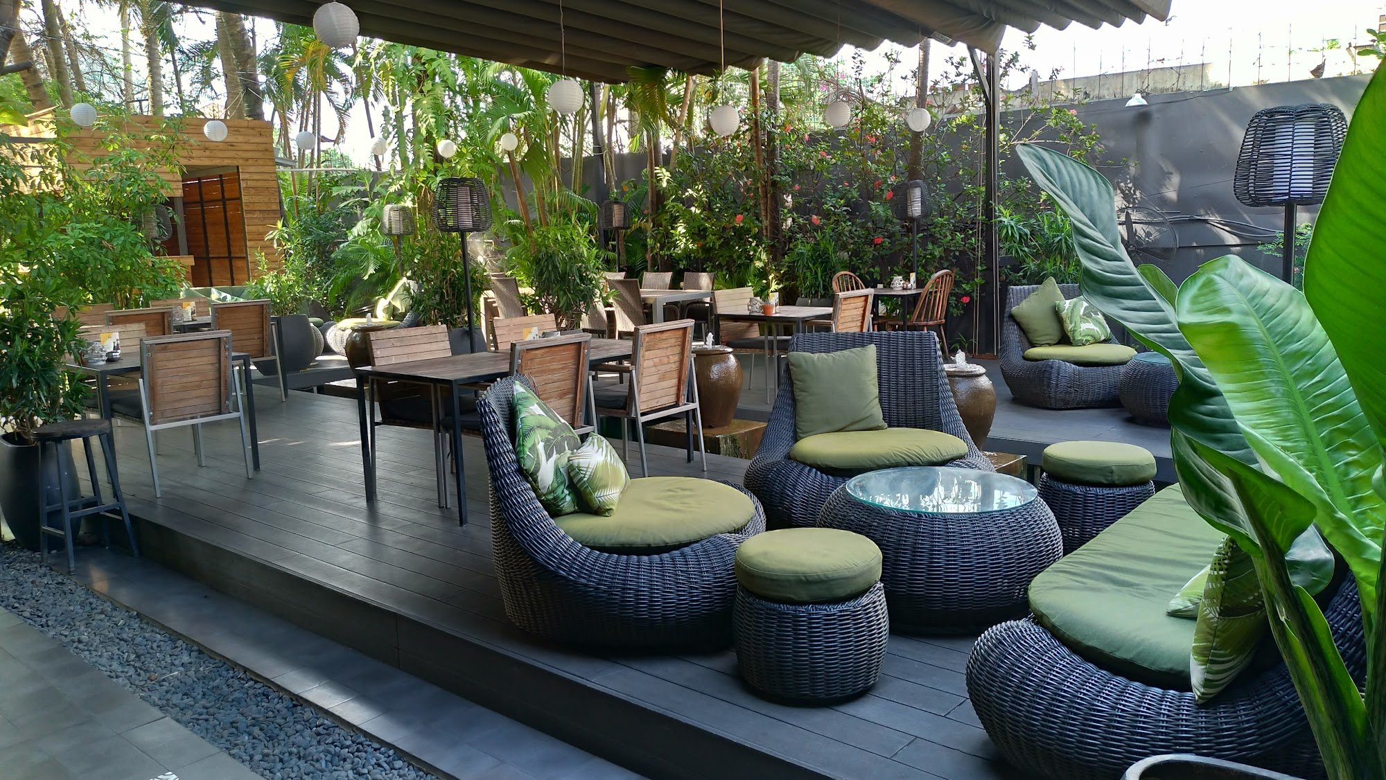 An outdoor seating area of a venue in Thao Dien, Saigon, featuring wicker chairs with green cushions, wooden tables, hanging white lanterns, lush trop
