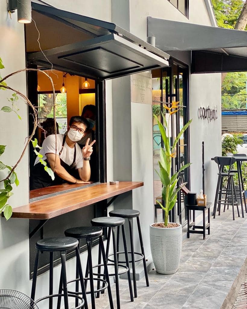 The image shows an exterior view of a modern coffee shop or bar named 'ecourtyard'. A person wearing glasses and a face mask is leaning on the counter