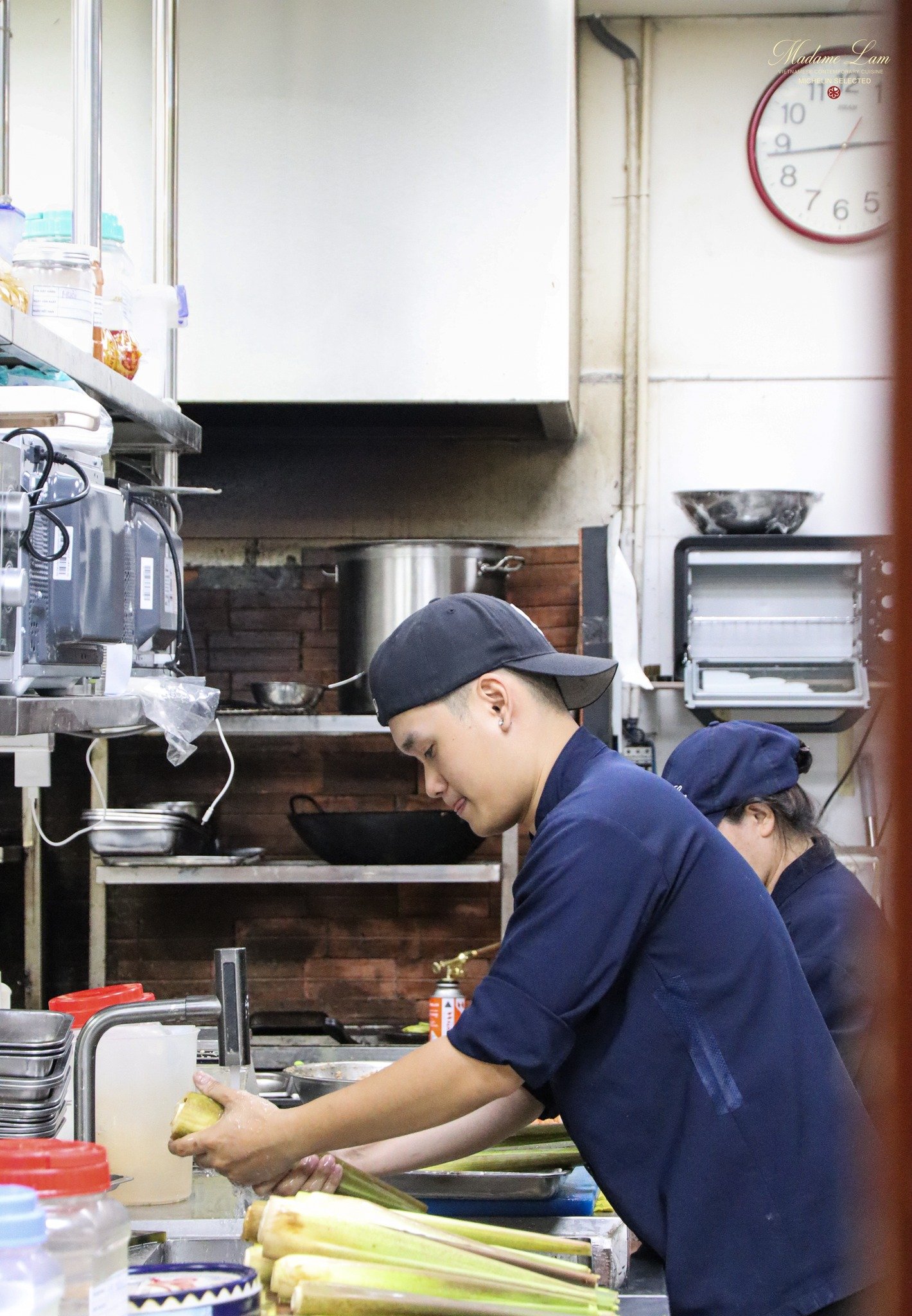 A person in a blue uniform is washing green stalks of vegetables or herbs at a commercial kitchen counter.