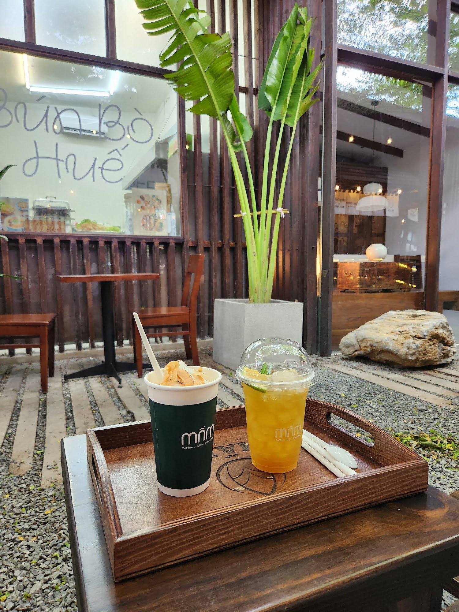 The image shows an outdoor seating area of a cafe named 'Bun Bo Hue' in Thao Dien, Saigon. The setting includes wooden furniture and green plants whic