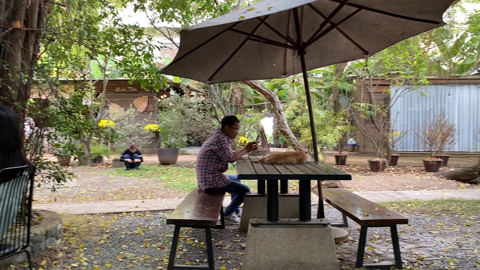 The image depicts an outdoor seating area in a garden-like setting at Thao Dien, Saigon. A man is seated on one side of the table with his legs crosse