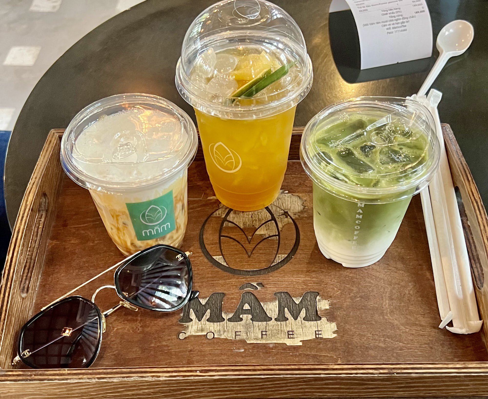 A wooden tray holds three beverages and a pair of sunglasses on top of it.