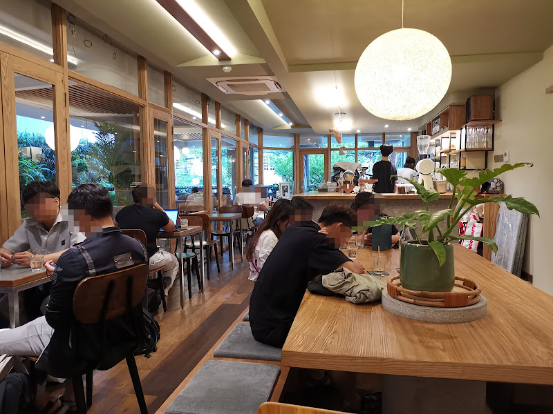 The image depicts the interior of a modern café in Thao Dien, Saigon. It features wooden tables and chairs, large glass windows with greenery visible 