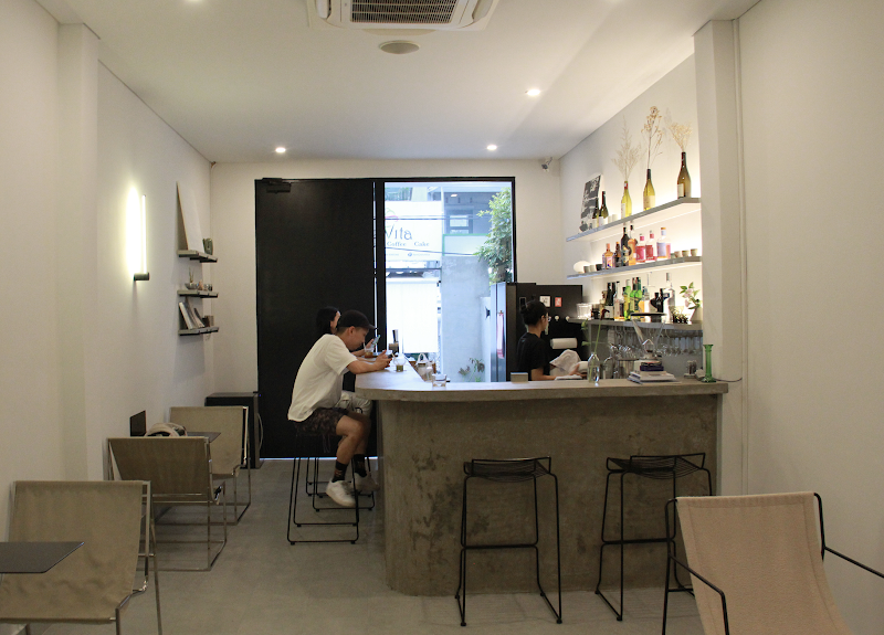 The image depicts the interior of a modern café or bar in Thao Dien, Saigon. It features a concrete bar counter with black metal stools, shelves displ