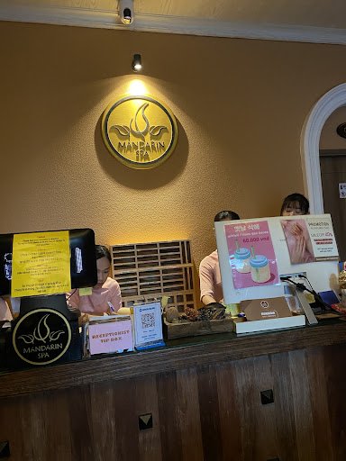 The image shows an interior view of a spa reception area named 'Mandarin Spa'. There are two staff members behind the counter, with various items such