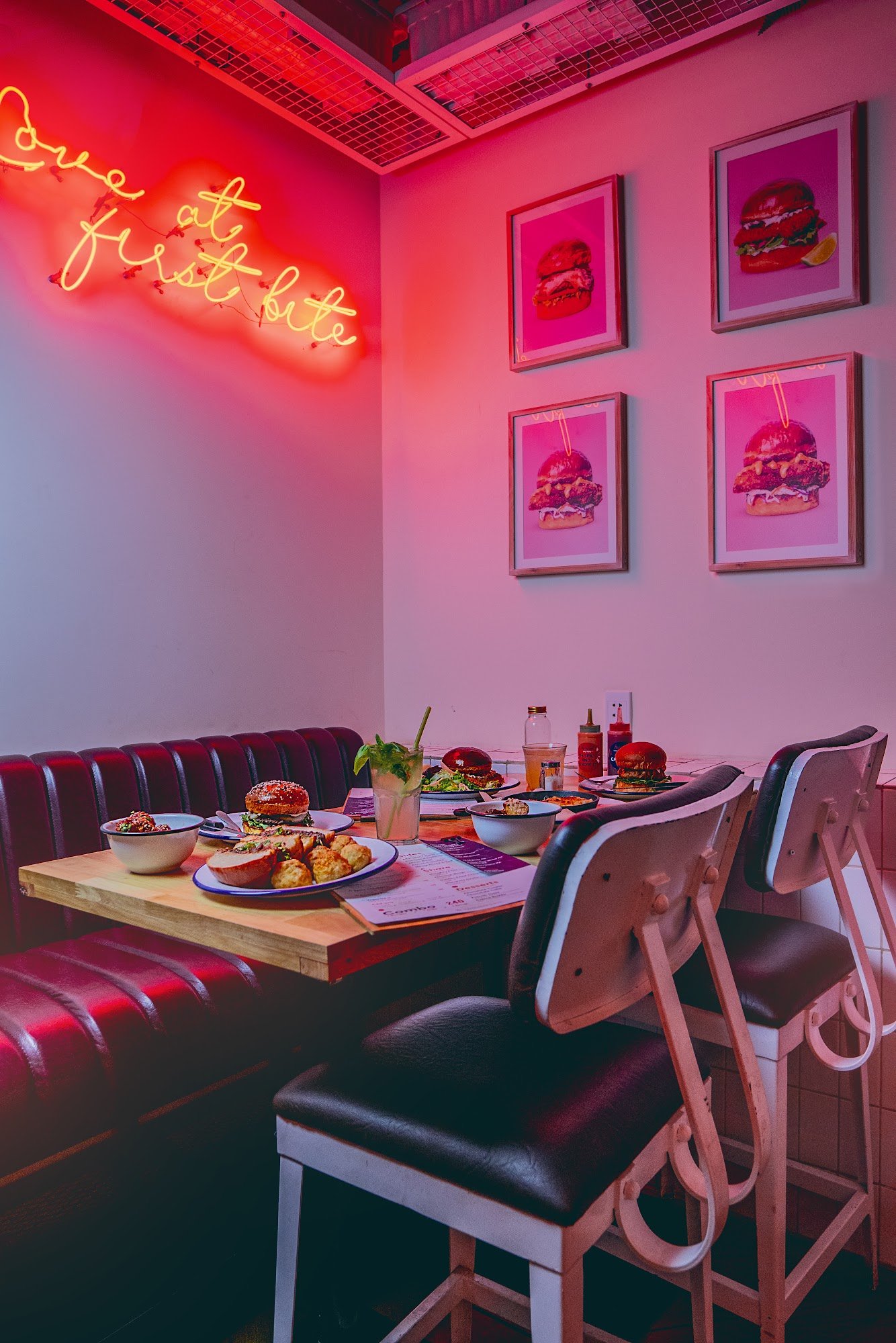 A vibrant restaurant interior in Thao Dien, Saigon, featuring a neon sign reading "love at first bite," pink-hued walls with framed burger artwork, a 