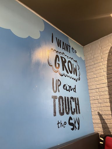 A motivational quote on a wall in an interior space that reads 'I WANT to GROW UP and TOUCH the SKY'. The background is blue with white clouds, sugges