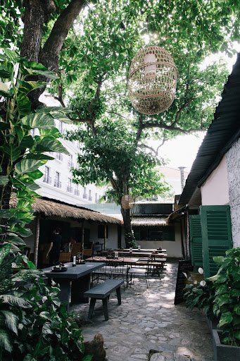 The image depicts an outdoor seating area of a restaurant or café in Thao Dien, Saigon. The space is shaded by large trees and features rustic wooden